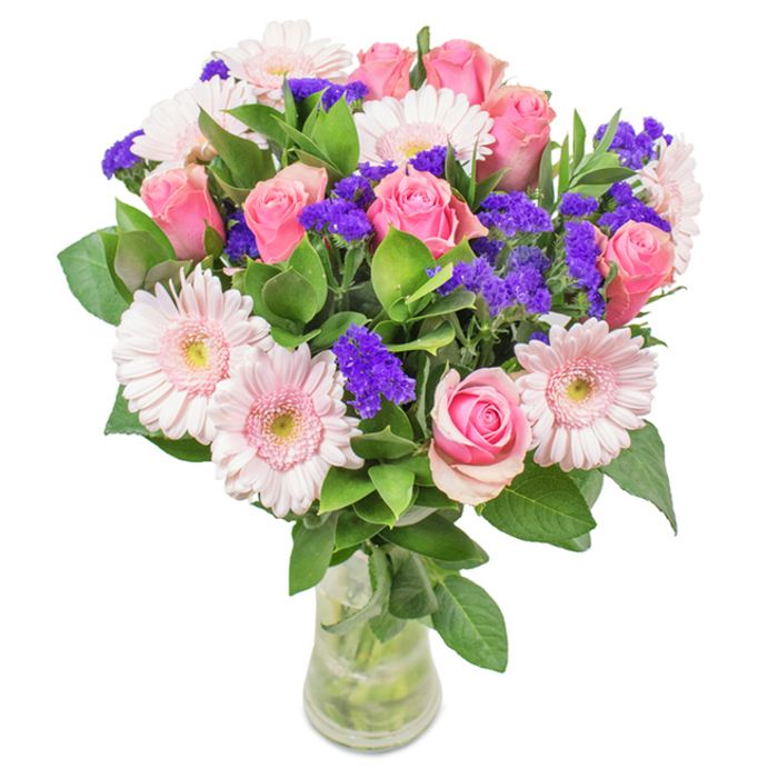 A fresh arrangement from Uxbridge Florist rests within a sparkling clear glass vase, backed by soft, natural light that enhances the palette's gentle warmth. At its heart are about seven to nine pink roses-some tightly closed, others opening in elegant spirals-showcasing a timeless floral form cherished throughout Uxbridge. Pale pink gerbera daisies can be counted among five to seven, their large, round heads and subtle creamy centers radiating outwards, recalling the open blooms seen in Fassnidge Park come early May. Scattered among the larger flowers are clusters of rich purple statice, each stem adding vibrant texture and contrast, much like the diverse stalls lining Uxbridge High Street. Deep emerald foliage forms a full, fresh base, supporting the blooms and hinting at the scent of morning dew on local lawns. The see-through vase reveals healthy green stems and crystal-clear water, highlighting the bouquet's just-picked energy and care in preparation. This arrangement makes a beautiful choice for birthdays, anniversaries, Mother's Day, or as a thoughtful accent for anyone living or working along the fringes of Hillingdon and Harefield. Every component signals joy, celebration, and genuine affection, inviting recipient and onlooker alike to pause, appreciate, and feel uplifted.